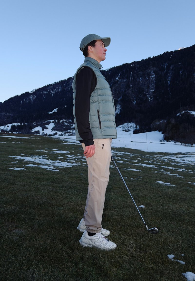 Νέος άνδρας με καπέλο και γιλέκο στέκεται σε χιονισμένο γήπεδο γκολφ κρατώντας ένα μπαστούνι γκολφ, με βουνά και καθαρό ουρανό στο παρασκήνιο.