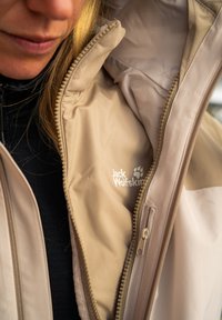 Beige takki, jossa on vetoketjullinen suljenta ja pehmeä tekstuuri. Siinä on näkyvä logo ja kontrastivärinen vuori, suunniteltu ulkokäyttöön.