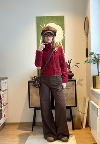 Femme portant une veste rouge, un pantalon marron à fines rayures, un chapeau beige et des lunettes, debout à l'intérieur devant une œuvre d'art verte et un meuble en bois.