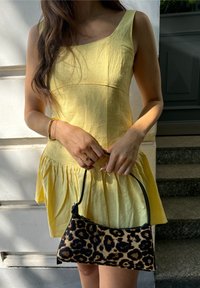 Frau in ärmellosem gelbem Kleid hält eine kleine Handtasche mit Leopardenmuster vor einer weißen Wand und steinernen Stufen.