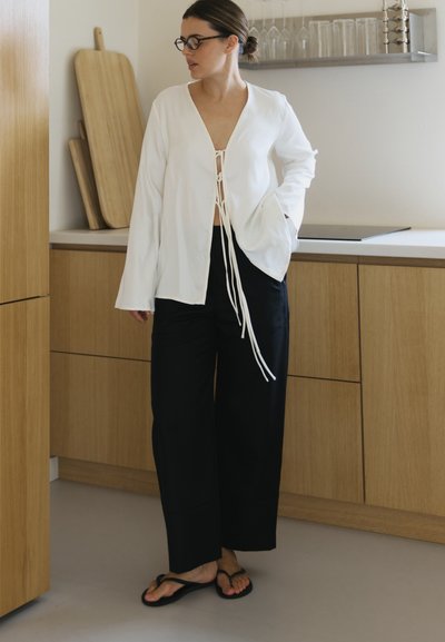 Mujer con pantalones negros, blusa blanca de frente abierto con cuerdas colgantes, chanclas negras y gafas, de pie en una cocina con armarios de madera.