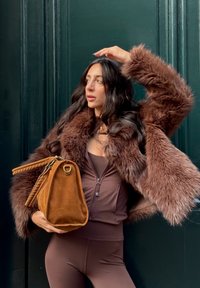 Brauner Pelzmantel mit strukturiertem Finish, kombiniert mit einem braunen Reißverschluss-Oberteil und hält eine braune Wildleder-Tasche mit gewebtem Akzent und goldenen Beschlägen.