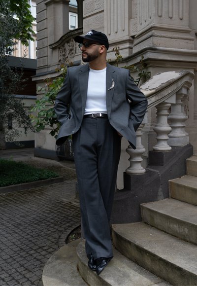 Hombre con barba que lleva un traje gris, camisa blanca, gorra negra y gafas de sol, de pie con las manos en los bolsillos en las escaleras de piedra de un edificio ornamentado.
