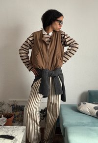 Personne portant des lunettes noires, un gilet marron à fermeture éclair, une chemise rayée à manches longues et un pantalon, avec un pull gris noué autour de la taille, à l'intérieur près des meubles.