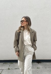 Blazer oversize beige indossato sopra una t-shirt bianca, abbinato a pantaloni a righe crema. Gli accessori includono orecchini a cerchio dorati e grandi occhiali da sole neri.