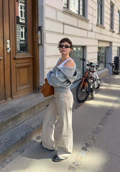 Persona con gafas de sol, suéter gris con los hombros descubiertos, camiseta blanca y pantalones anchos beige, de pie en una acera de la ciudad cerca de bicicletas.