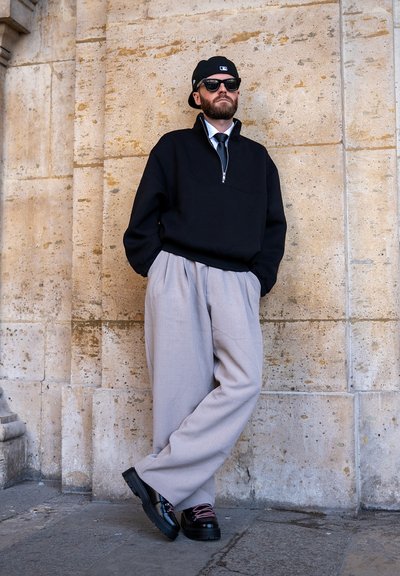 Hombre con gorra negra, gafas de sol, jersey negro, pantalones anchos gris claro y zapatos negros, de pie contra una pared de piedra beige con las manos en los bolsillos.