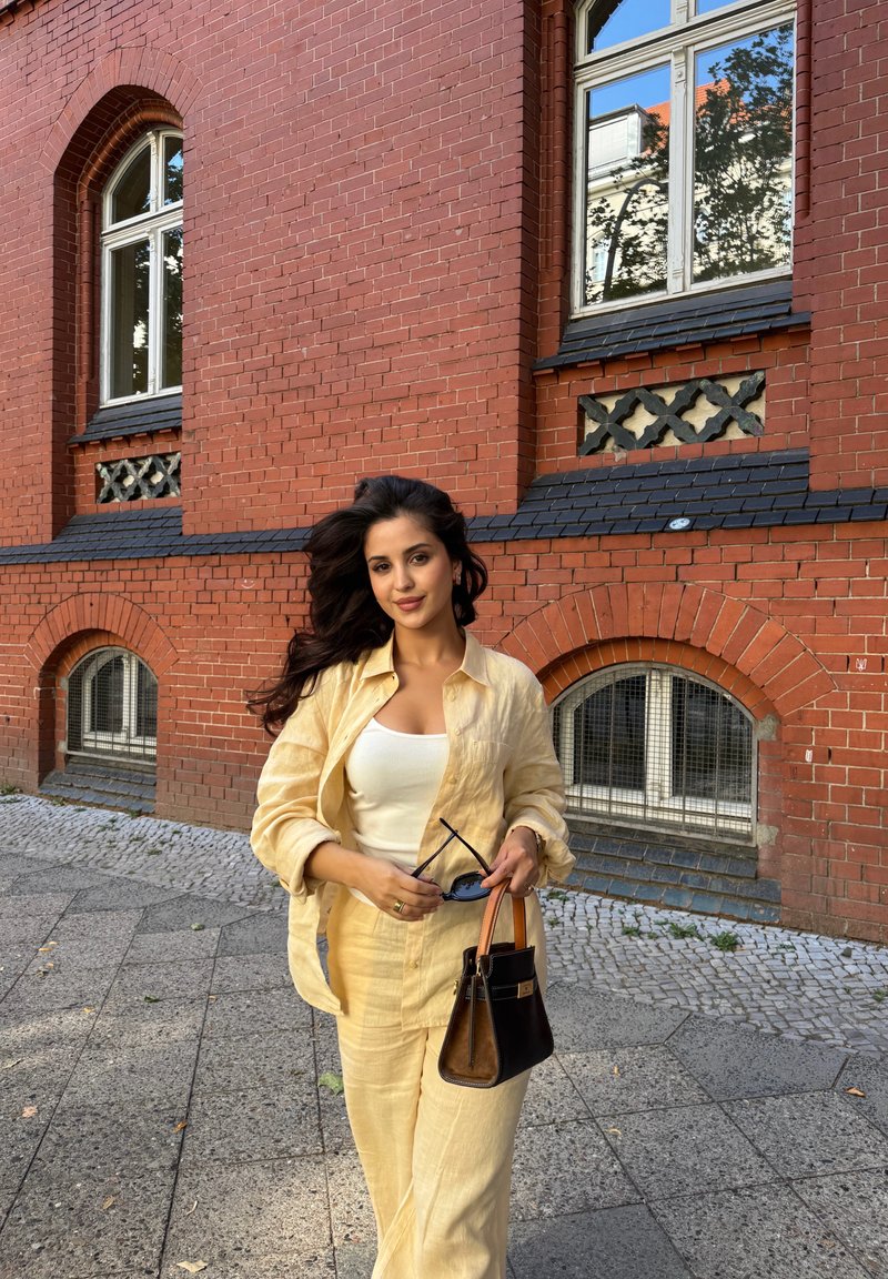 Femme en tenue jaune clair tenant des lunettes de soleil et un sac à main marron, debout sur le trottoir devant un bâtiment en briques rouges avec des fenêtres en arc.