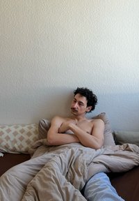 Κλινοσκεπάσματα σε μπεζ απόχρωση, συμπεριλαμβανομένου ενός παπλώματος και μαξιλαριού, τοποθετημένα πάνω σε υφασμάτινο καφέ στρώμα. Άτομο με σγουρά μαλλιά ξεκουράζεται στον χώρο.