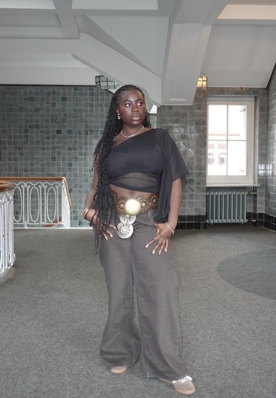 Top negro transparente de una manga, pantalones anchos marrones y hebilla redonda de cinturón de latón. Trenzas largas, maquillaje natural y zapatos neutros.