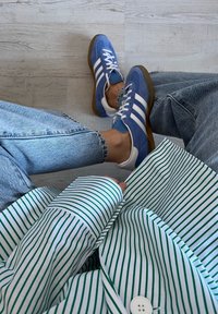 Μπλε σουέτ αθλητικά παπούτσια με λευκές ρίγες και καφέ σόλες, συνδυασμένα με λευκό πουκάμισο με πράσινες κατακόρυφες ρίγες.