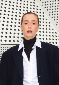 Svart oversize blazer över en vit skjorta, lager på lager med en figurnära svart polotröja. Släta hudtoner och geometrisk mönstrad bakgrund.