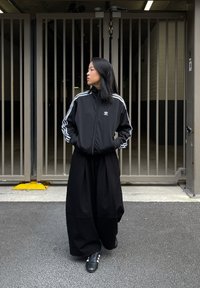 Nero giacca a zip con strisce bianche; pantaloni neri larghi; sneakers nere. Sullo sfondo ci sono cancelli di metallo e pavimentazione in cemento.