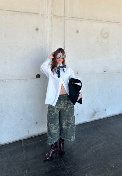 Camisa blanca con el frente abierto, bufanda negra, pantalones cortos cargo de camuflaje y tacones burgundy de punta. Gafas de sol reflectantes y chaqueta negra en la mano.