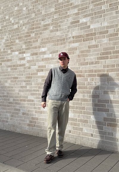 Joven con chaleco de punto gris con trenzas, camisa negra, pantalones claros y gorra burdeos, de pie frente a una pared de ladrillo clara al aire libre.