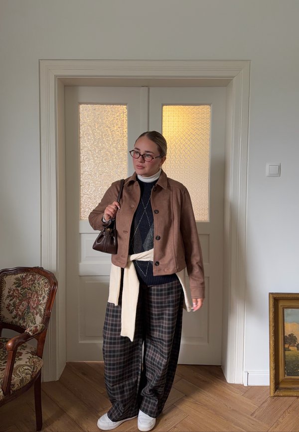 Mujer joven con gafas, chaqueta marrón, pantalones anchos de cuadros y zapatillas blancas está de pie dentro de una habitación cerca de una puerta sosteniendo un bolso marrón sobre su hombro.