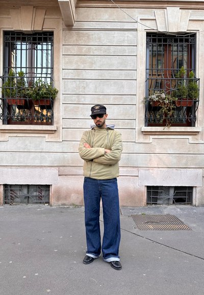 Chaqueta de color verde oliva con cremallera y capucha gris, pantalones vaqueros azules holgados con puños doblados, zapatos negros y gorra negra con acentos blancos. Fondo neutro.