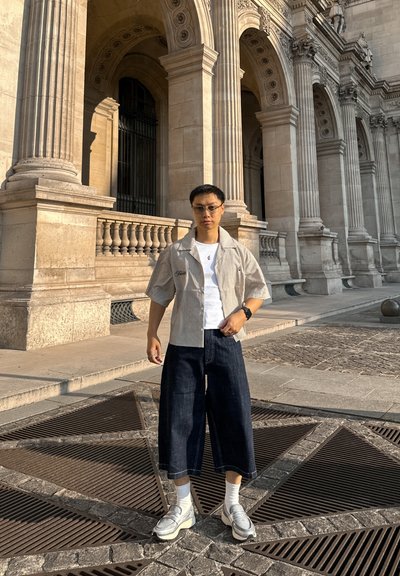 Camisa de rayas de manga corta, camiseta blanca debajo, pantalones de mezclilla oscuros de pierna ancha, calcetines blancos y zapatillas deportivas. El fondo presenta columnas de piedra.