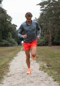 Szara, długorękawowa koszulka sportowa, pomarańczowe szorty i dopasowane buty do biegania. Biegnąc po piaszczystej ścieżce otoczonej drzewami.