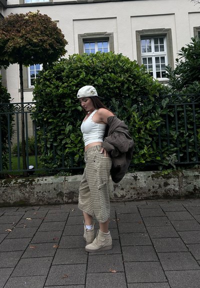 Mujer joven con gorra blanca, top blanco de tirantes, pantalones anchos de cuadros y zapatillas beige, sosteniendo una chaqueta marrón, de pie en la acera cerca de arbustos verdes.