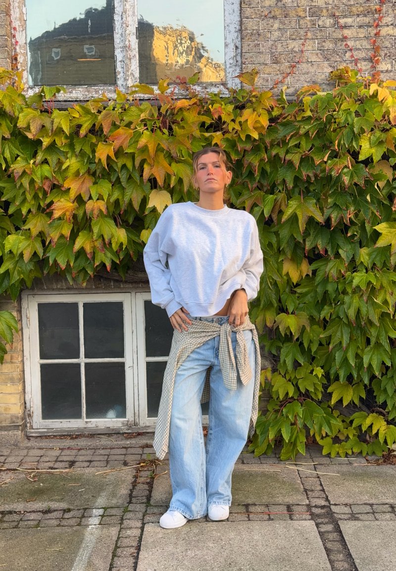 Une femme pose en sweat-shirt gris, en jean ample et en baskets blanches devant un mur de briques avec des fenêtres, couvert de lierre.