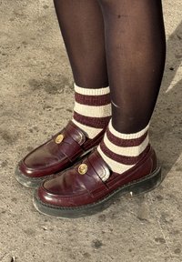 Mocassins en similicuir bordeaux avec des ornements dorés décoratifs, associés à des chaussettes rayées crème et bordeaux, portées sur des collants noirs.