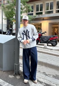 Grijze gebreide trui met donkerblauw NY-logo, gecombineerd met wijdvallende donkerblauwe spijkerbroek en lichtgekleurde instapschoenen. Witte pet met NY-logo.