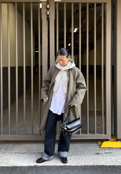 Persona con ropa en capas que lleva un abrigo gris, sudadera con capucha y camisa blanca, de pie frente a una alta puerta metálica, sosteniendo un bolso negro.