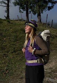Lila bolyhos mellény egy barna hosszú ujjú póló felett, bézs hátizsákkal felszerelve. Csíkos kötött sapka és napszemüveg teszi teljessé a szabadtéri megjelenést.