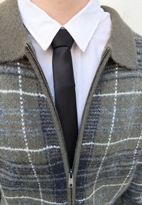 Veste en laine à fermeture éclair avec un motif à carreaux gris et noir, dotée d'un col et d'une fermeture éclair métallique, associée à une chemise blanche et une cravate noire.