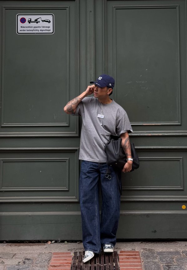 Mand med tatoveringer iført kasket, grå t-shirt og jeans står foran grønne dobbeltdøre og holder en sort taske, mens han justerer sine øretelefoner.