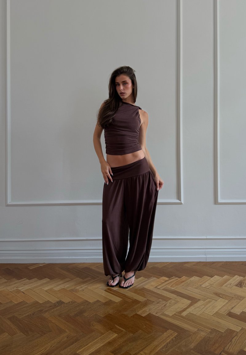 Femme debout à l'intérieur portant un haut sans manches marron foncé et un pantalon ample assorti sur un parquet en bois.