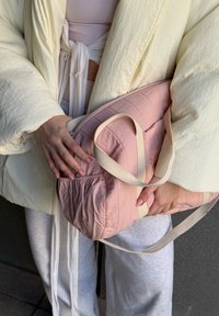 Personne portant une veste crème et un pantalon clair tient un sac matelassé rose avec des sangles beige sur un fond sombre.