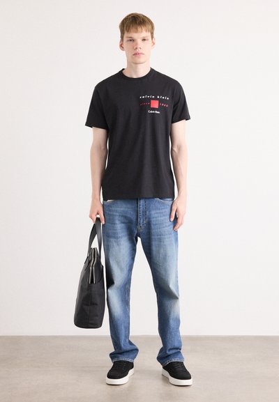 Camiseta de algodón negra con un logo rojo y blanco, combinada con unos vaqueros de mezclilla azules y zapatillas negras, sosteniendo una bolsa tote negra.