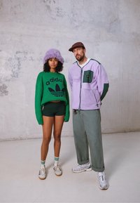 Sweater de malha verde com o logótipo "adidas", chapéu fofinho roxo, calções pretos, ténis brancos. Homem com uma polar lilás com fecho, detalhes verdes, calças cinzentas.