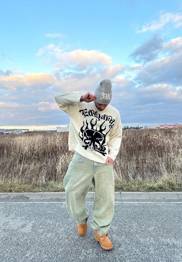 Pull en crème tricoté avec un design de crâne noir et de flammes, associé à un pantalon vert menthe ample et des bottes chevilles marron. Bonnet rayé gris.