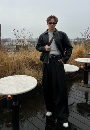 Chaqueta de cuero negra, suéter gris de canalé, pantalones anchos negros, zapatillas blancas, gafas de sol redondas. Entorno al aire libre con césped y mesas.