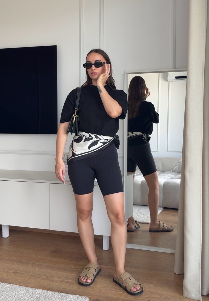 Femme portant des lunettes de soleil noires, un t-shirt noir, un cuissard noir, une écharpe imprimée nouée autour de la taille, des sandales marron clair, debout à l'intérieur près d'un miroir.