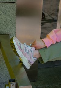 New Balance joggesko i lyserosa og hvit med mesh-paneler, fremtredende logo, teksturerte såler, brukt med grønne bukser på en metallbenk.