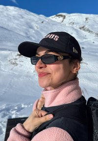 Femme portant une casquette noire et des lunettes de soleil, vêtue d'une polaire rose, dans un paysage montagneux enneigé sous un ciel bleu clair.