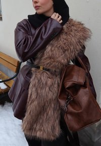 Femme ajustant une écharpe noire, portant une veste en cuir marron avec un gilet en fausse fourrure et portant un grand sac en cuir marron à l'extérieur dans un décor enneigé.