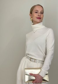 Jeune femme en col roulé crème et jupe plissée, tenant une pochette métallique dorée, portant des boucles d'oreilles dorées et un collier délicat.