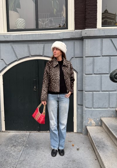 Chaqueta con manchas marrones y crema, suéter negro, vaqueros azul claro, zapatos negros, sosteniendo un bolso rojo con detalles en crema y marrón frente a un edificio.