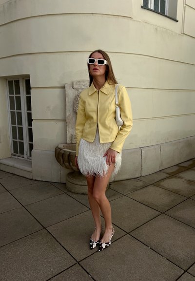 Mujer con chaqueta amarilla, falda blanca con plumas, tacones con estampado de vaca y gafas de sol blancas, de pie en una acera pavimentada cerca de una pared curva beige.