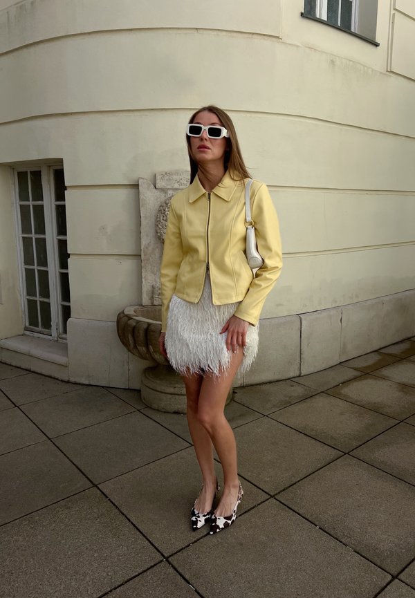 Femme portant une veste jaune, une jupe blanche plumeuse, des talons imprimés vache et des lunettes de soleil blanches, debout sur un trottoir pavé près d'un mur beige incurvé.