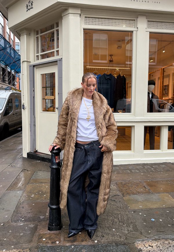 Frau in einem langen braunen Pelzmantel, weißem Hemd, lockeren dunklen Jeans und schwarzen spitzen Schuhen steht an einer nassen Straßenecke und lehnt sich an einen schwarzen Pfosten.