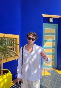 Camicia bianca con bottoni e due tasche, pantaloni beige, occhiali da sole neri. Ambientato su sfondi vivaci blu e giallo con dettagli in verde sulla porta.