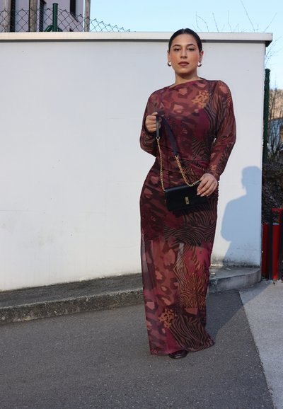 Vestido largo y transparente con un patrón de estampado animal en burdeos y marrón. La modelo sostiene un pequeño bolso negro con una correa de cadena dorada.
