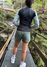 Crna i svijetloplava lagana jakna sa uzorkom trakama, zelene kratke hlače i bijele čarape. Osoba stoji na metalnoj rešetkastej cesti u šumi.