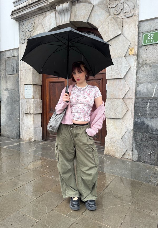 Jonge vrouw staat op nat trottoir met een zwarte paraplu, draagt een roze vest, een bloemenblouse, olijfgroene cargobroek en zwarte plateauzolen.